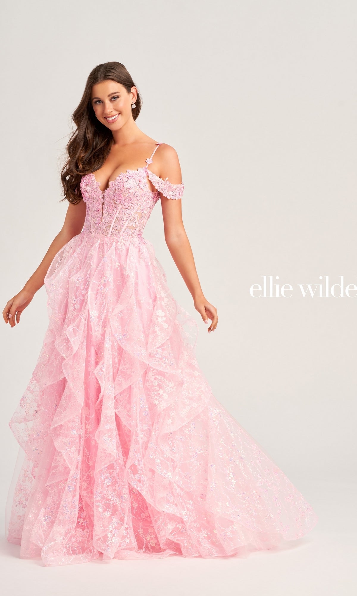 A woman wears the Ellie Wilde Ruffled Long Prom Ball Gown EW35218 in light pink, featuring off-shoulder ruffles, floral details, and beaded lace appliques, standing against a plain background with the designer's name visible on the dress.